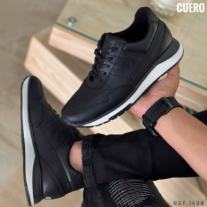 Zapato deportivo de cuero negro para caballero con suela blanca, mostrado en mano y pie con pantalón oscuro.